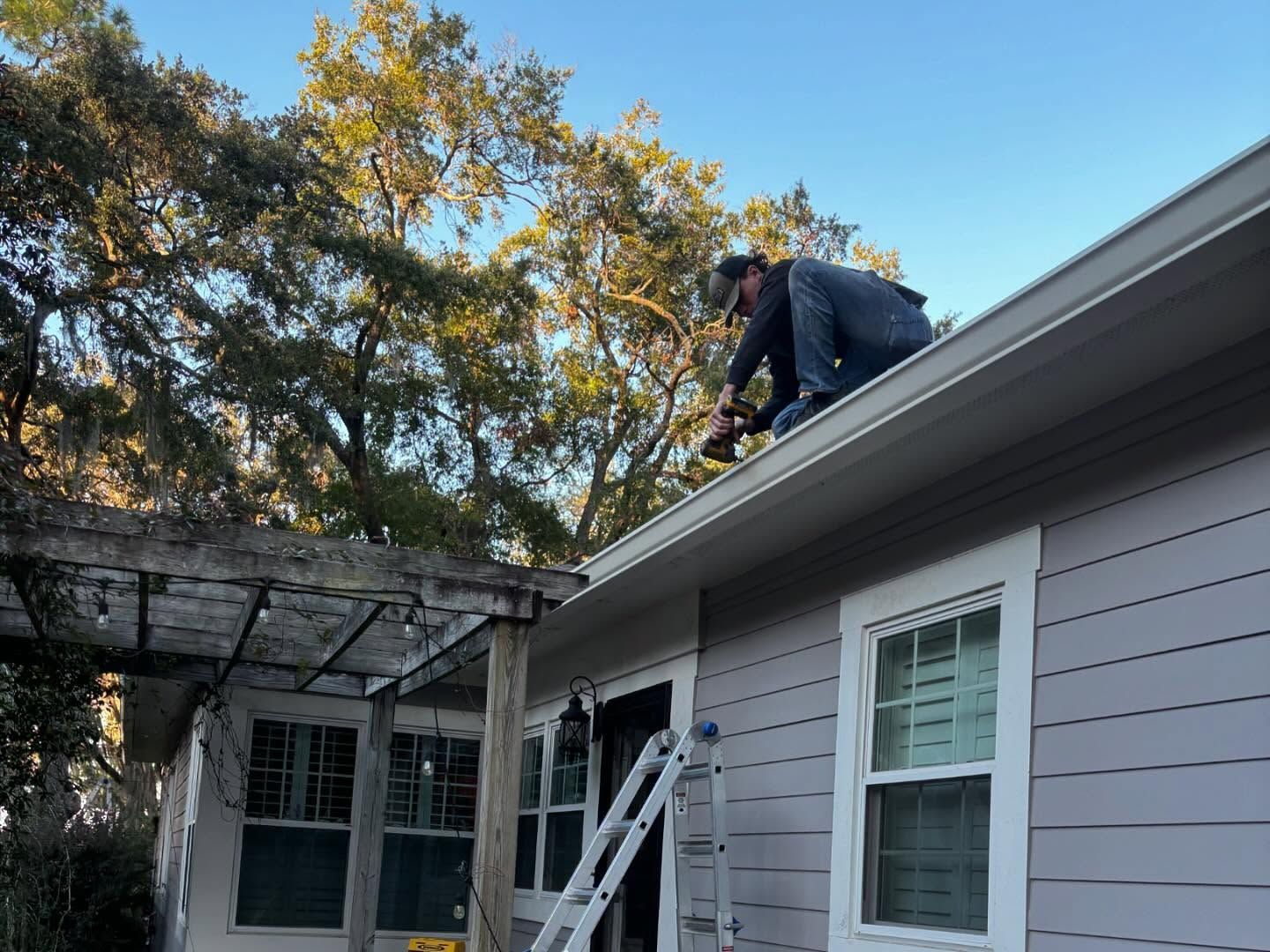 A person wearing a dark shirt and jeans works on the roof of a grey house next to a wooden pergola and a ladder.