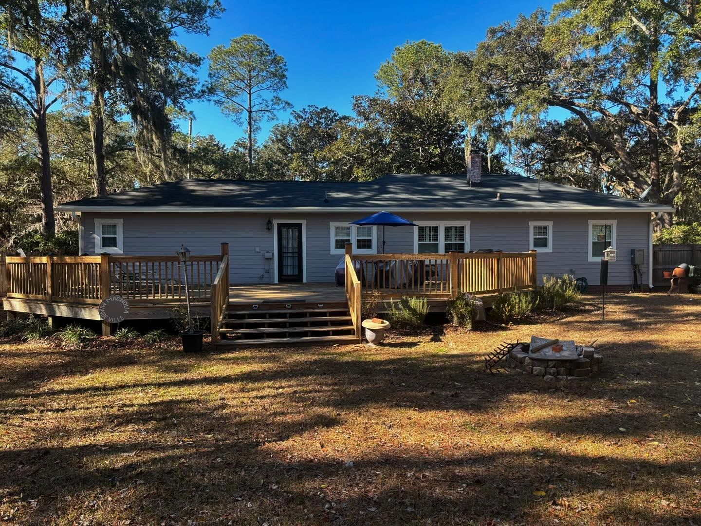 Gray single-story house with a large wooden deck in a wooded backyard under a clear blue sky.
