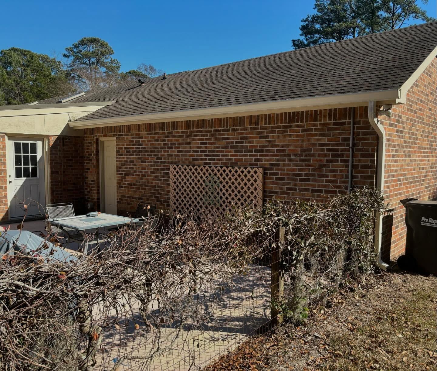 A brick house exterior with a patio, trellis, outdoor table, and a small fence in a backyard setting under a blue sky.