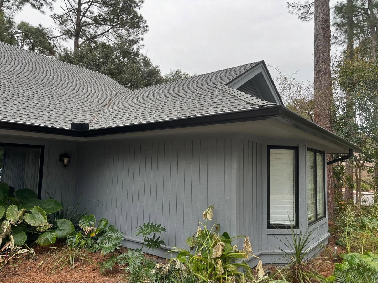 A gray, single-story house with board-and-batten siding, a dark shingled roof, and bay windows set among trees.