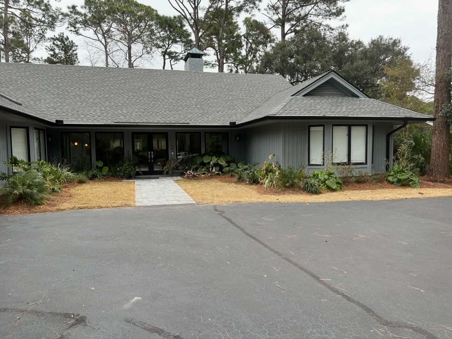 A grey one-story building with a shingled roof, glass double doors, and surrounding pine trees and bushes.