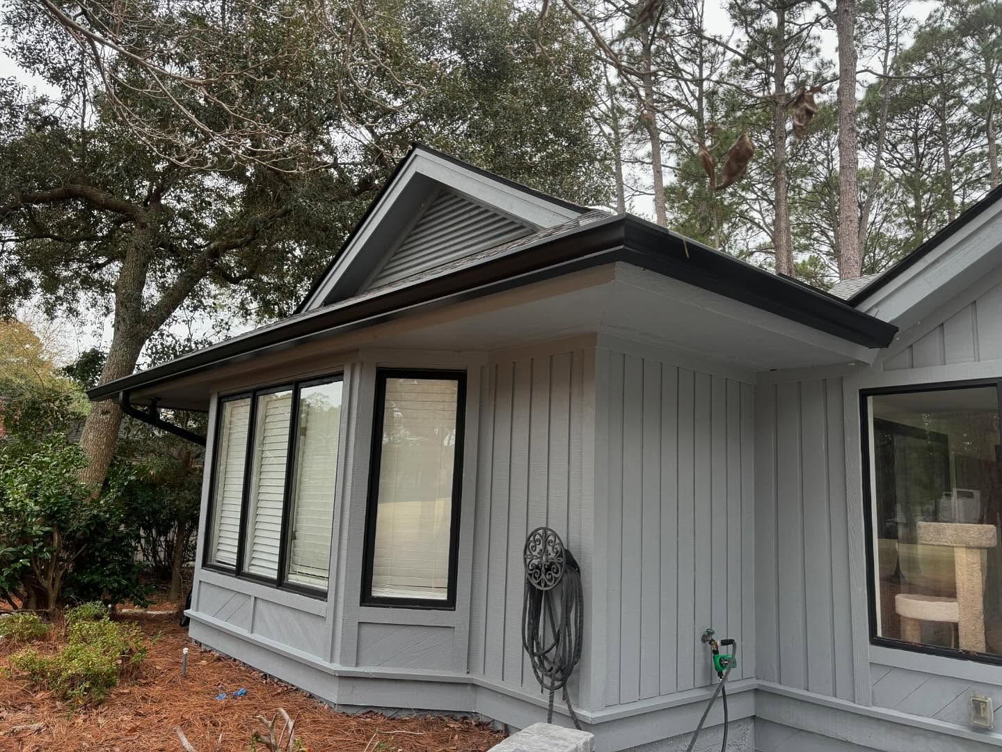A light gray house with vertical siding and black window trim features a bay window section under a gabled roof.