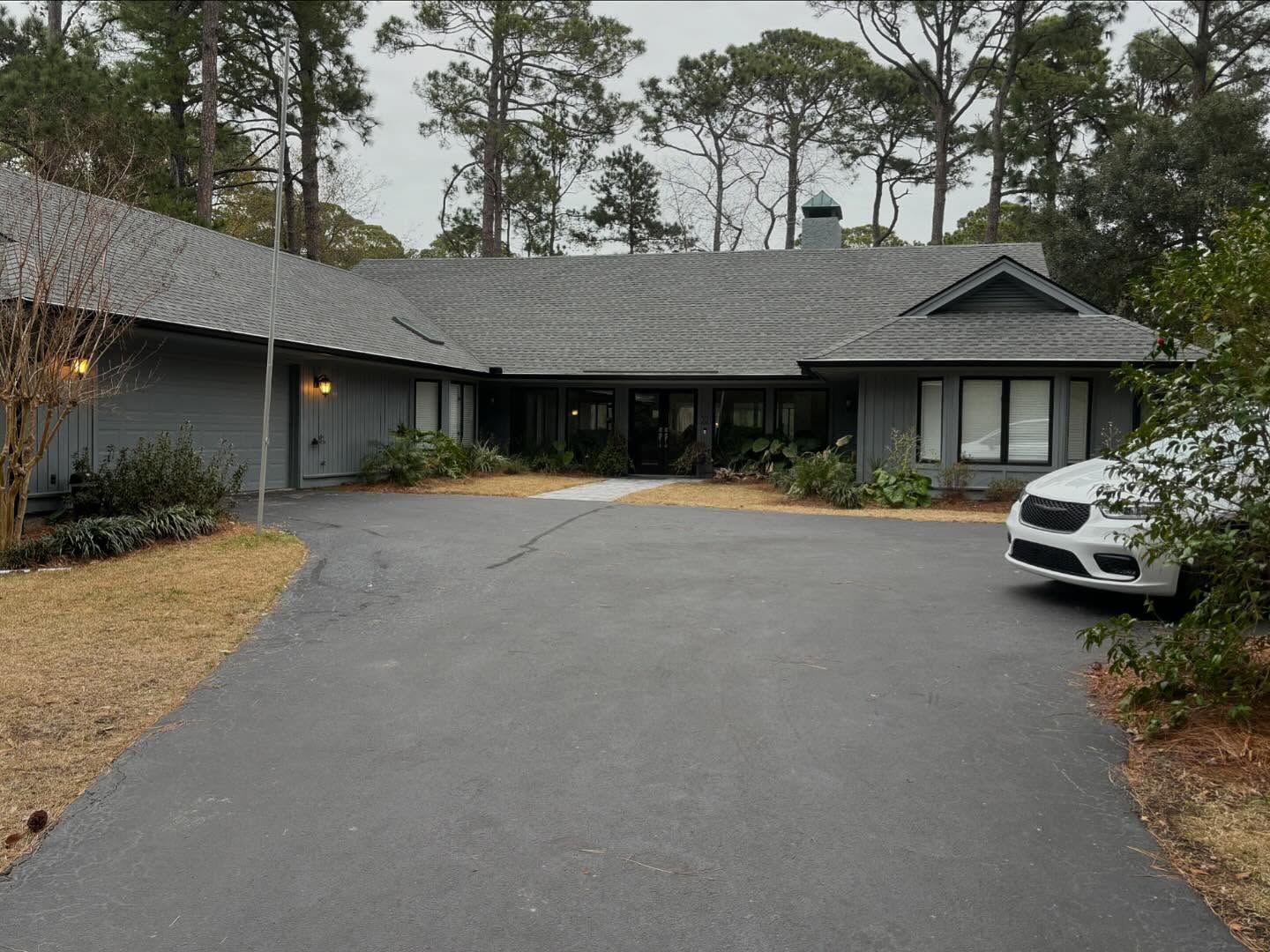 A single-story gray house with a dark roof and chimney sits behind a long paved driveway under a cloudy sky.