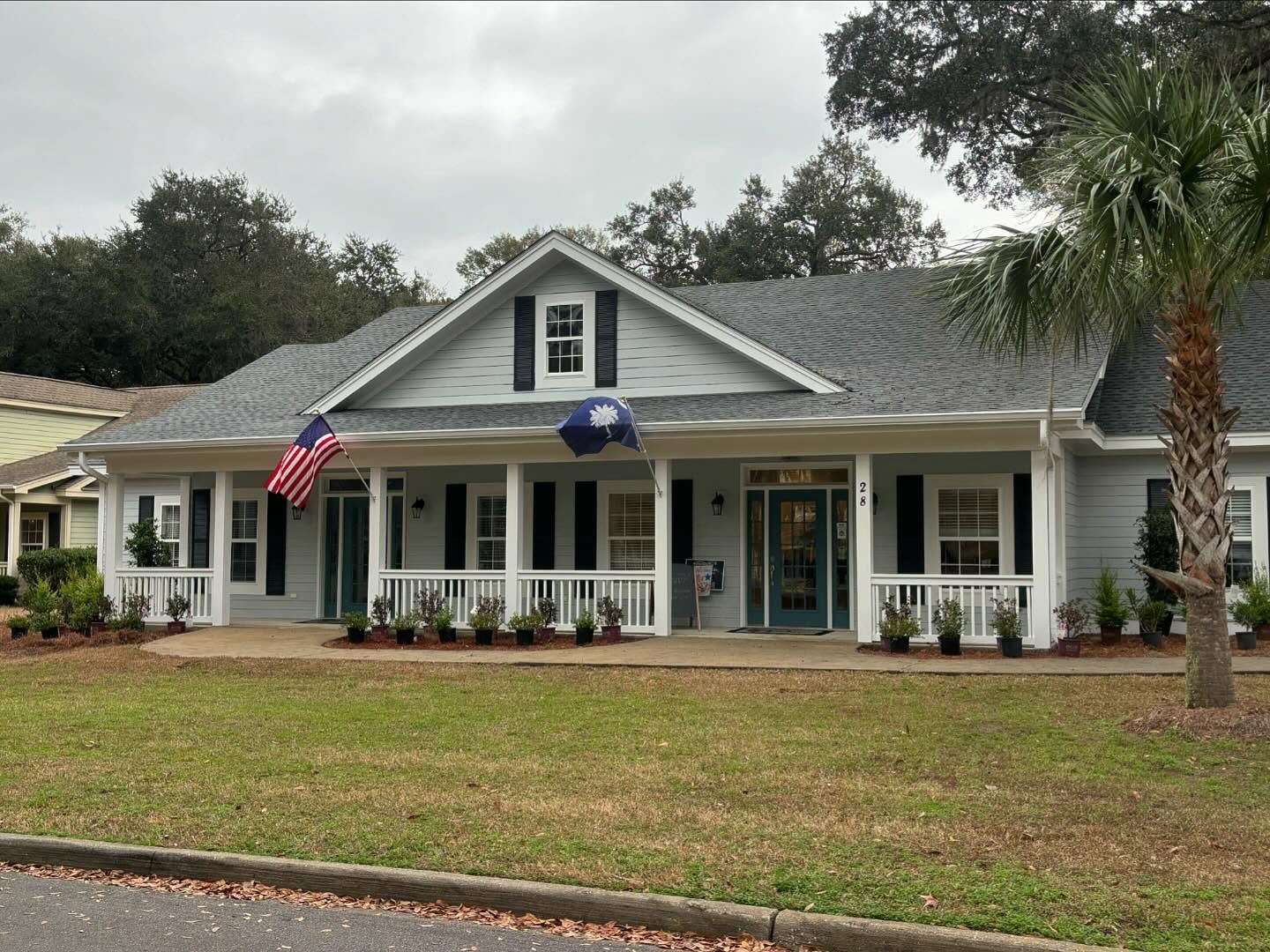 A one-story gray house with a full-width front porch, black shutters, and American and South Carolina flags.