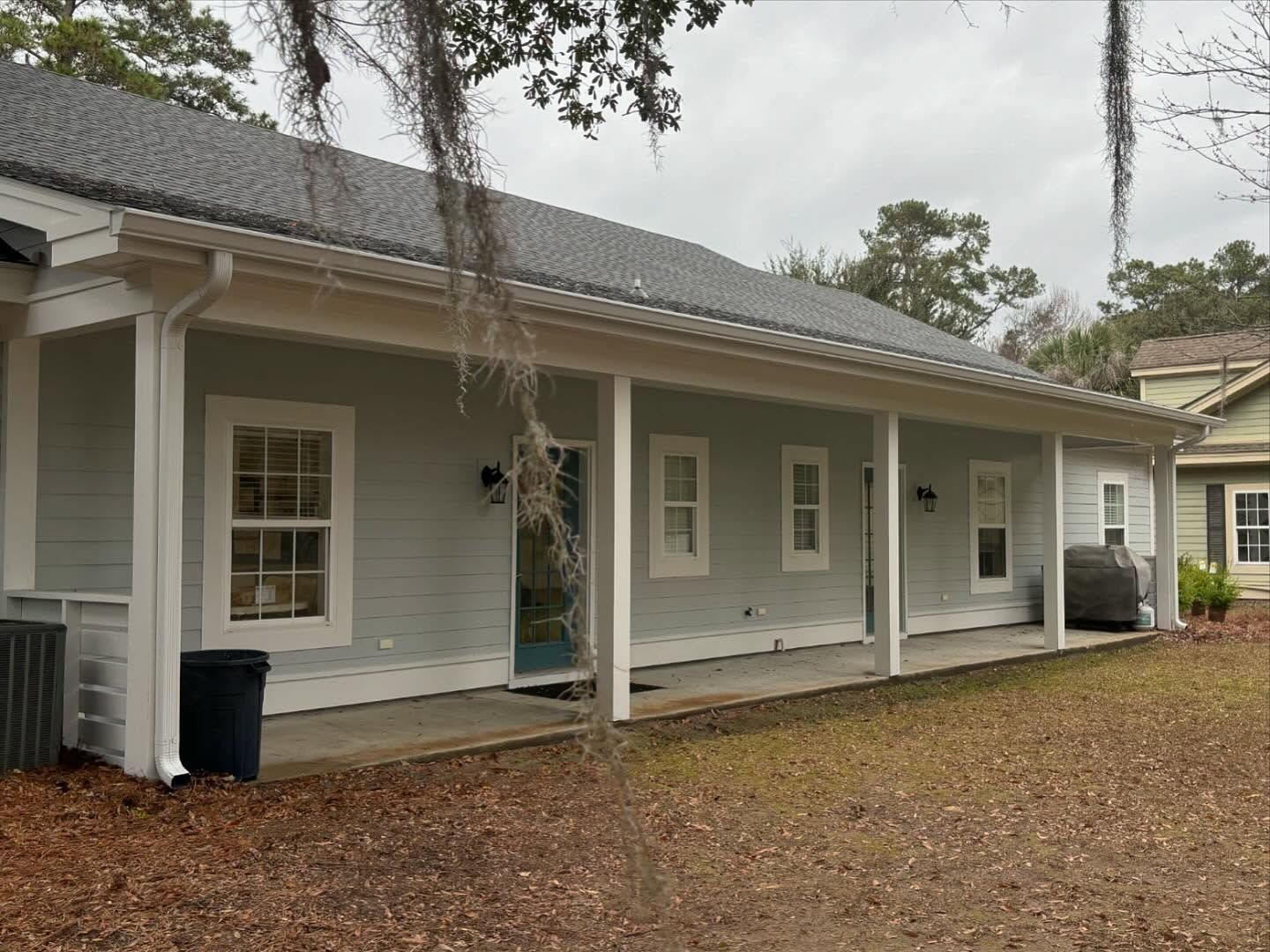 A light grey, single-story house with a long, white-pillared covered porch sits on a yard covered in fallen brown leaves.