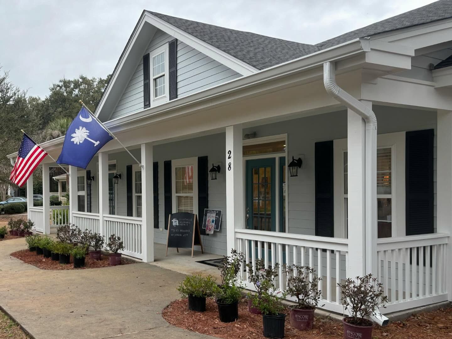 A light-colored house with a wraparound porch, South Carolina and American flags displayed, and potted plants in front.