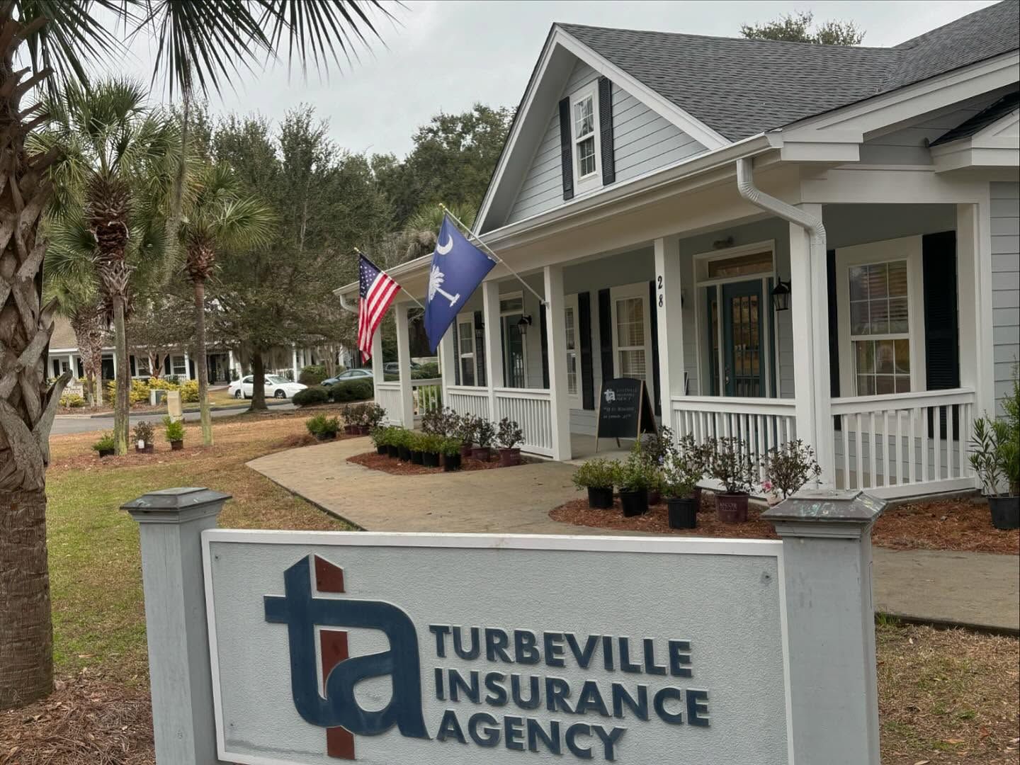 A sign for Turbeville Insurance Agency stands in front of a light gray building with a porch and American and state flags.