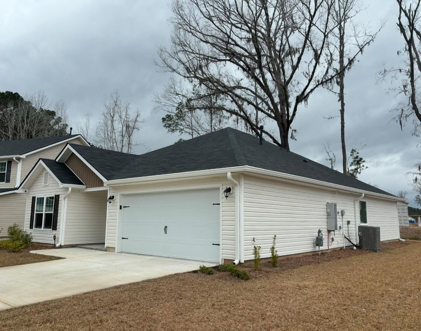 A single-story residential house with white vinyl siding, a dark roof, and a two-car garage under a cloudy sky.