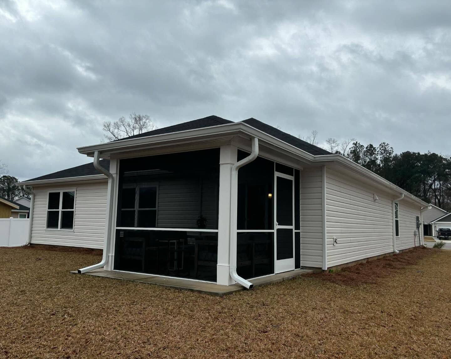 A white, one-story house with a black roof and a screened-in porch on a lawn under a cloudy sky.