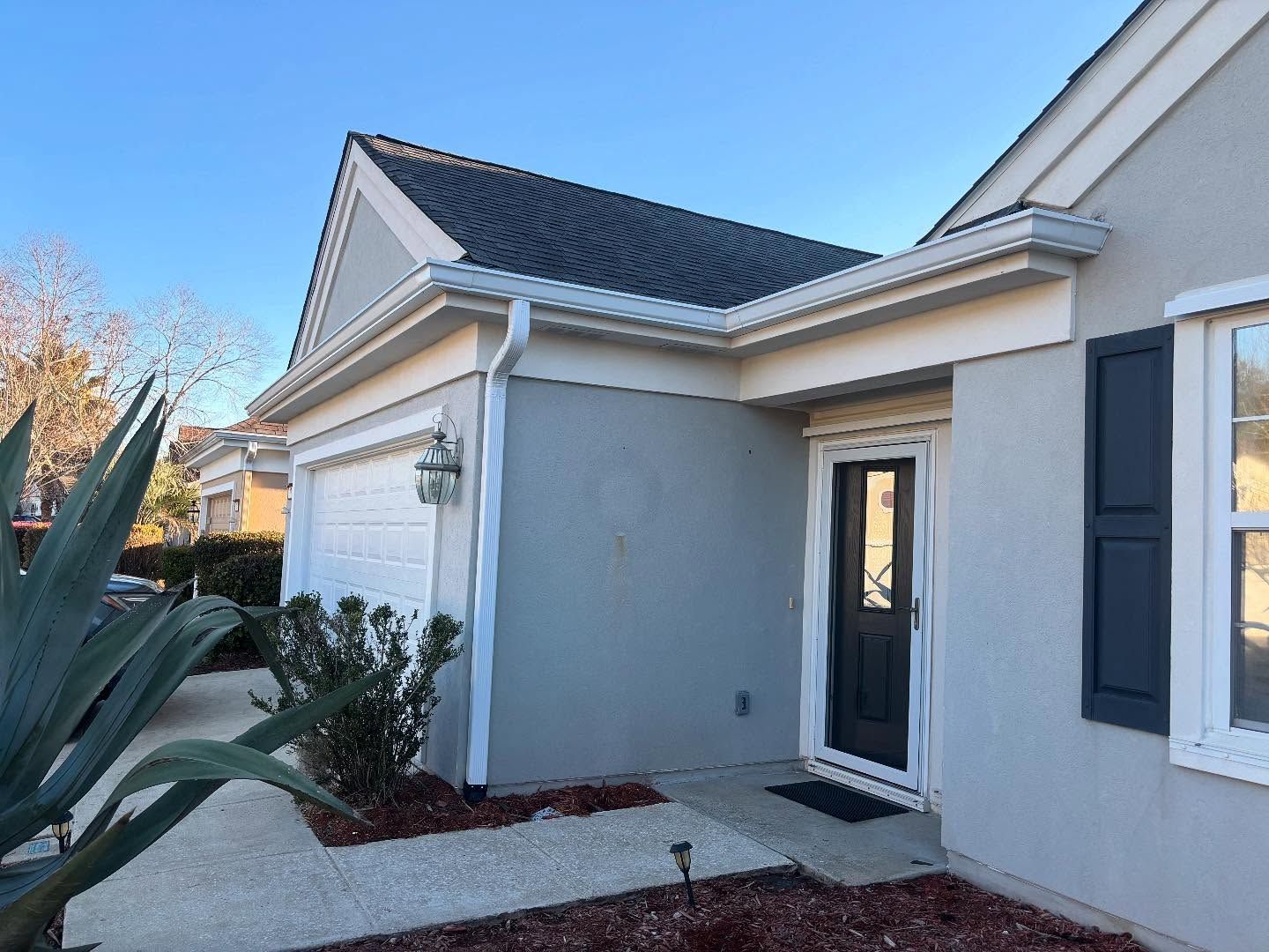A gray one-story house exterior featuring a white garage door, a black front door, and a dark shingled roof under blue sky.