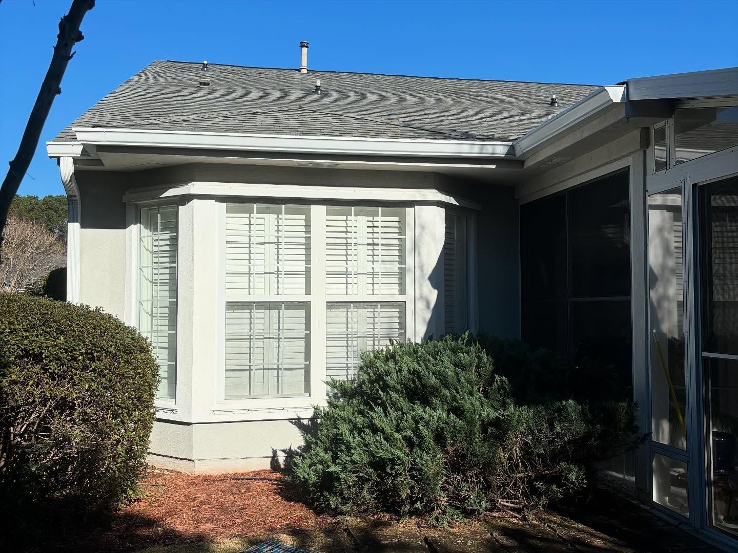 A bay window on a neutral-colored house with a shingled roof, surrounded by landscaping and a screened porch area.