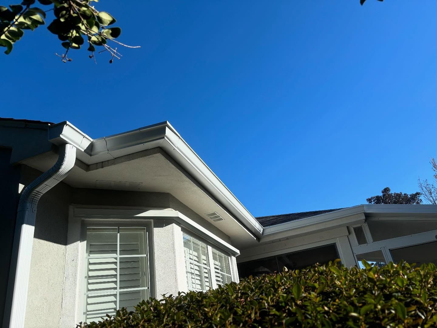 Low-angle view of a residential home exterior with a light-colored stucco wall, white trim, and a bush under a blue sky.
