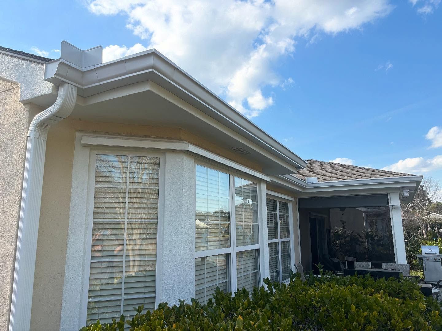 A low-angle view of a beige house exterior with white trim, gutters, shutters, and a covered patio area under a blue sky.