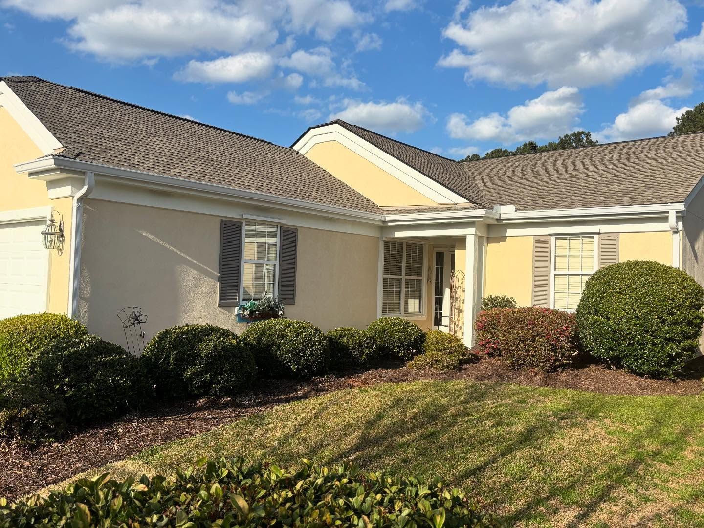 A single-story tan stucco home with a gray shingled roof, dark shutters, and manicured green hedges under a blue sky.