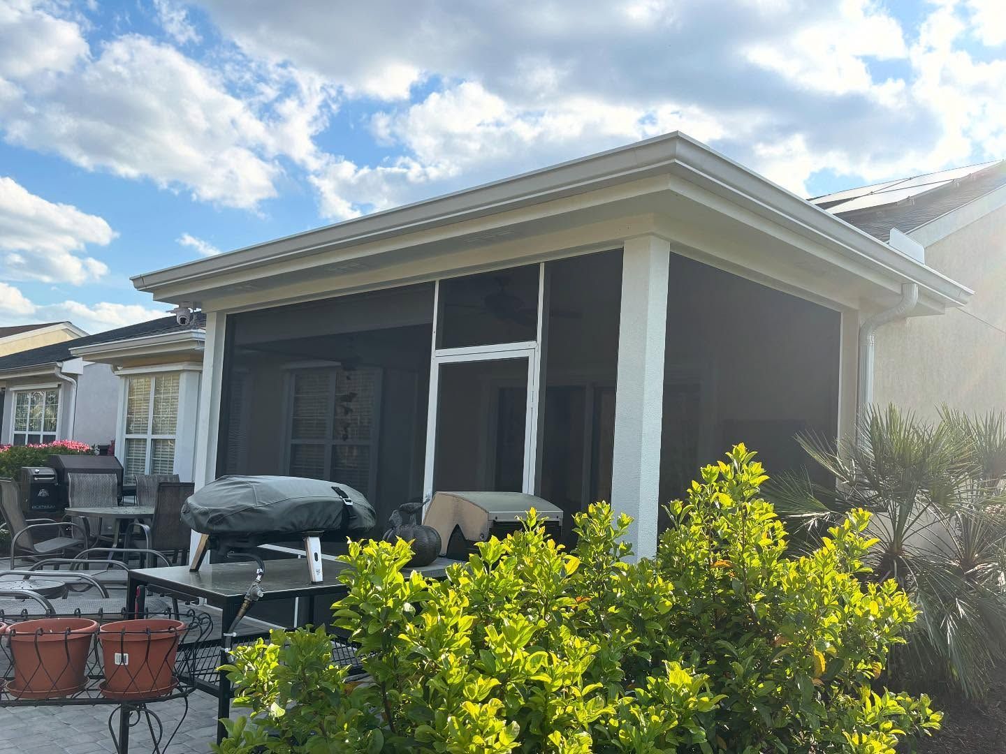 A covered outdoor patio with a screened enclosure, a table, and a grill, viewed from a sunny garden setting.