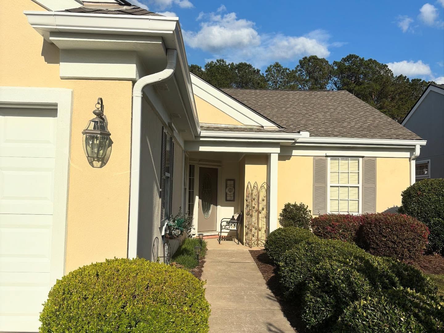 A tan suburban house with a paved walkway, a prominent front door, white trim, and trimmed green bushes under a blue sky.