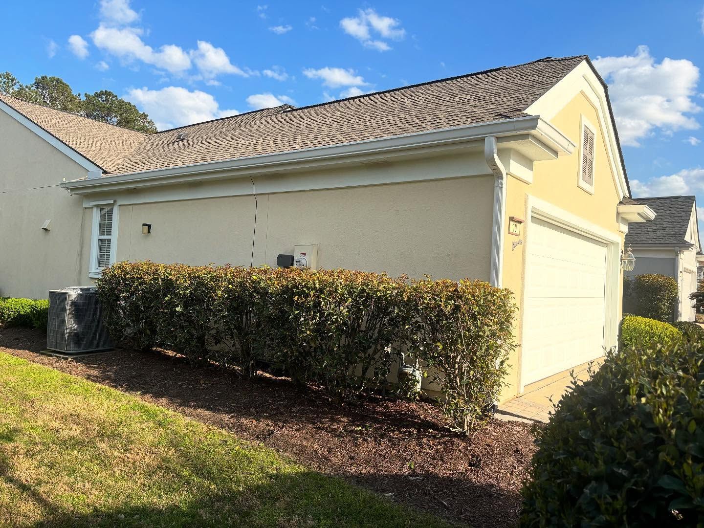 Side view of a beige suburban home with a garage, a trimmed hedge, and a grass lawn under a blue, cloudy sky.