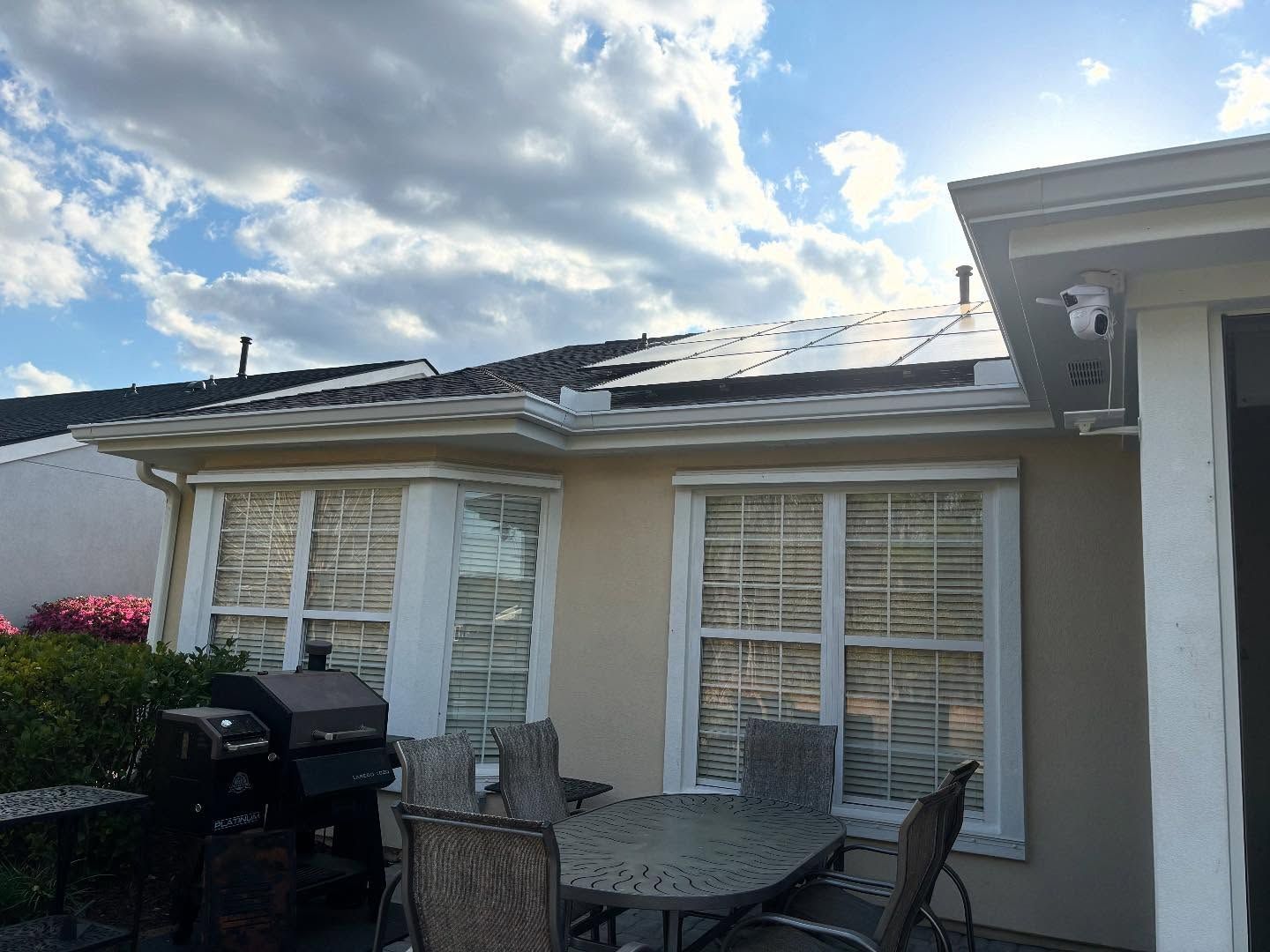 Patio furniture and a grill sit outside a home with a roof featuring solar panels against a cloudy blue sky.