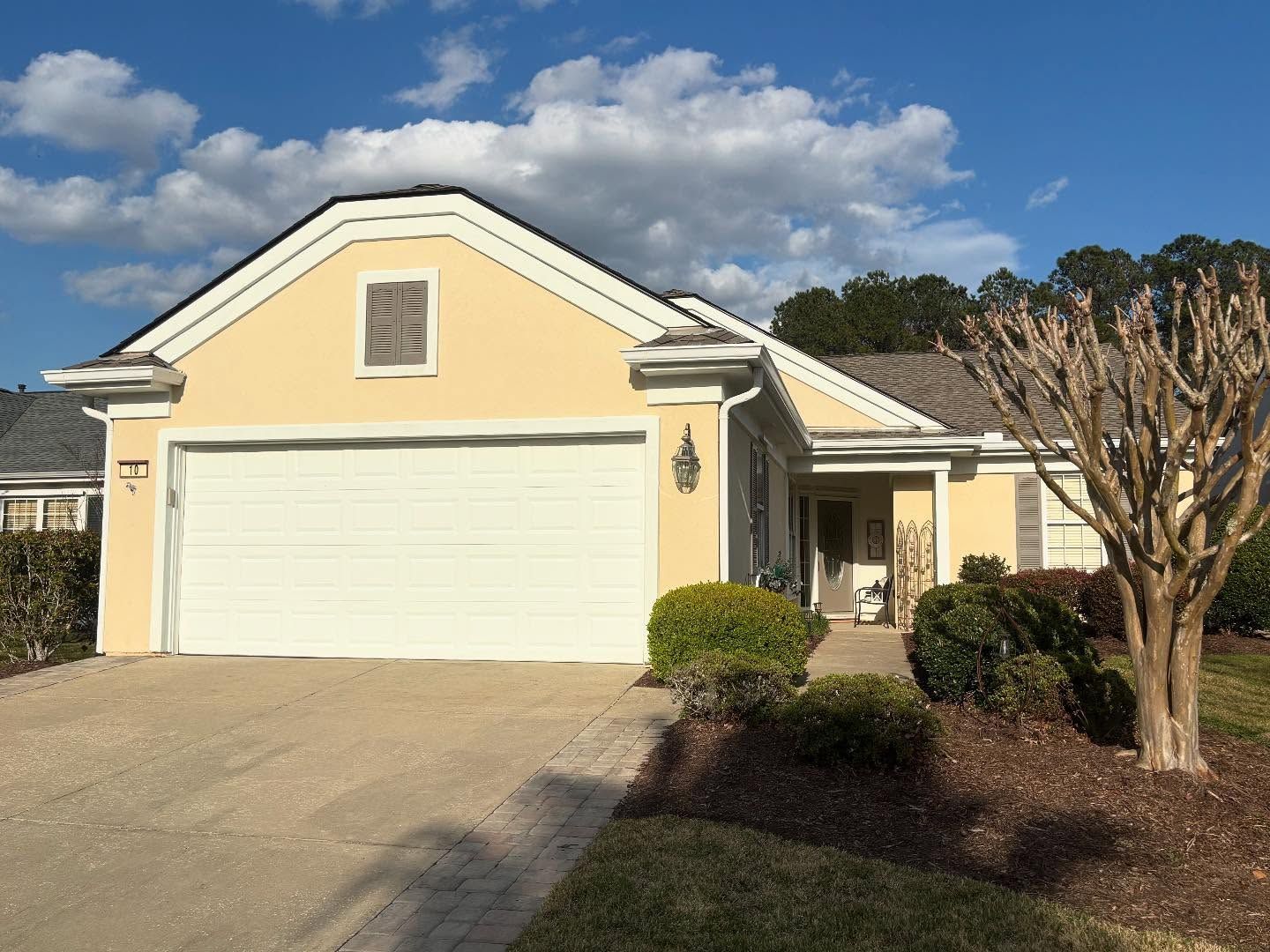 A pale yellow, single-story house with a white garage door, concrete driveway, and a bare tree in the front yard.