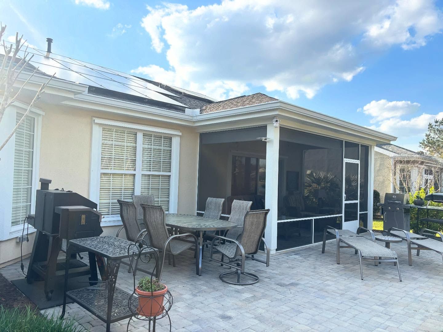 A sunlit patio featuring a grill, a table with four chairs, and a screened-in porch against a beige house.