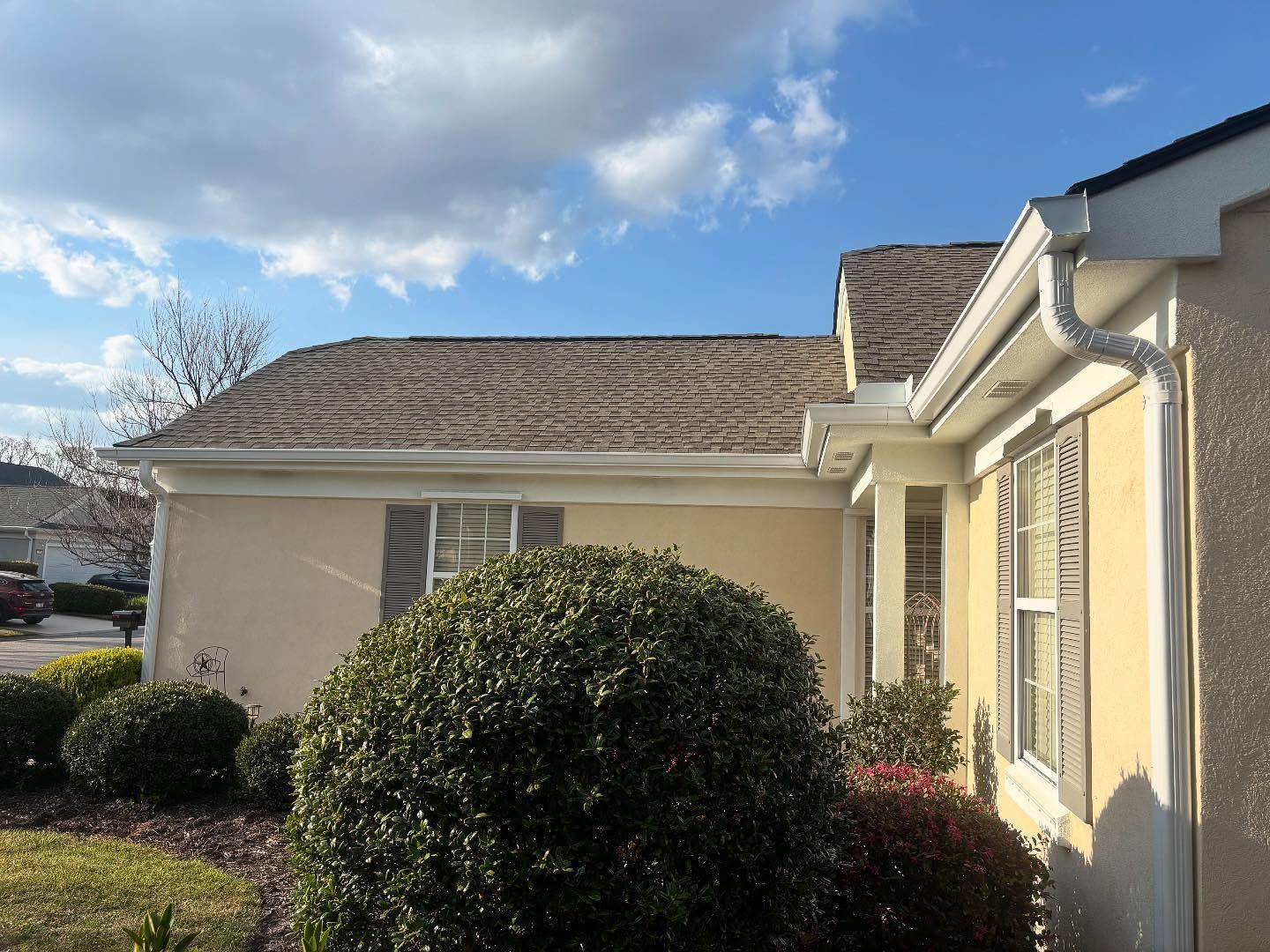 A beige, one-story house with a shingled roof, white gutters, and a large green bush in the front yard under a blue sky.