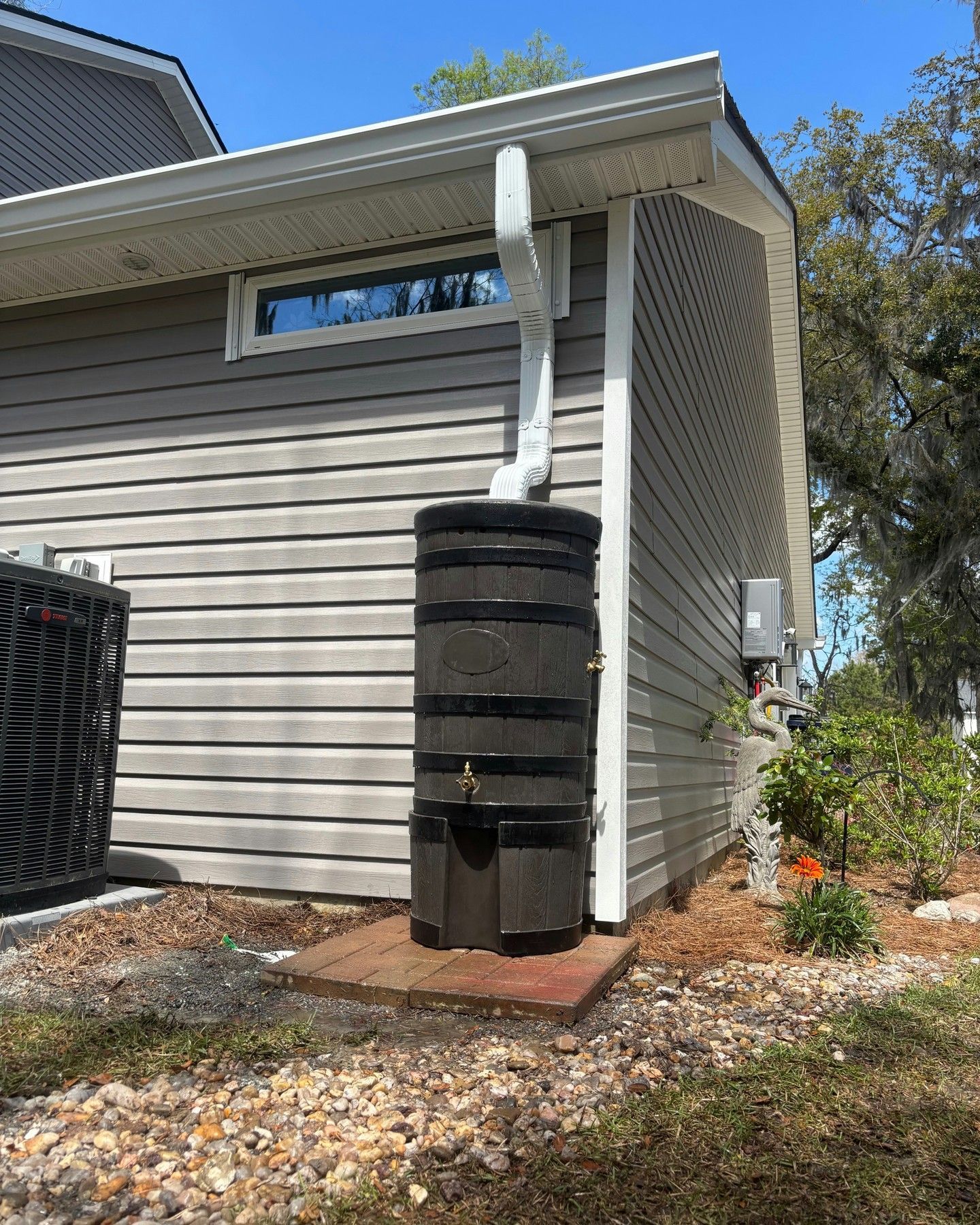 A dark brown rain barrel stands on a brick base against a grey vinyl-sided house wall, connected to a white downspout.