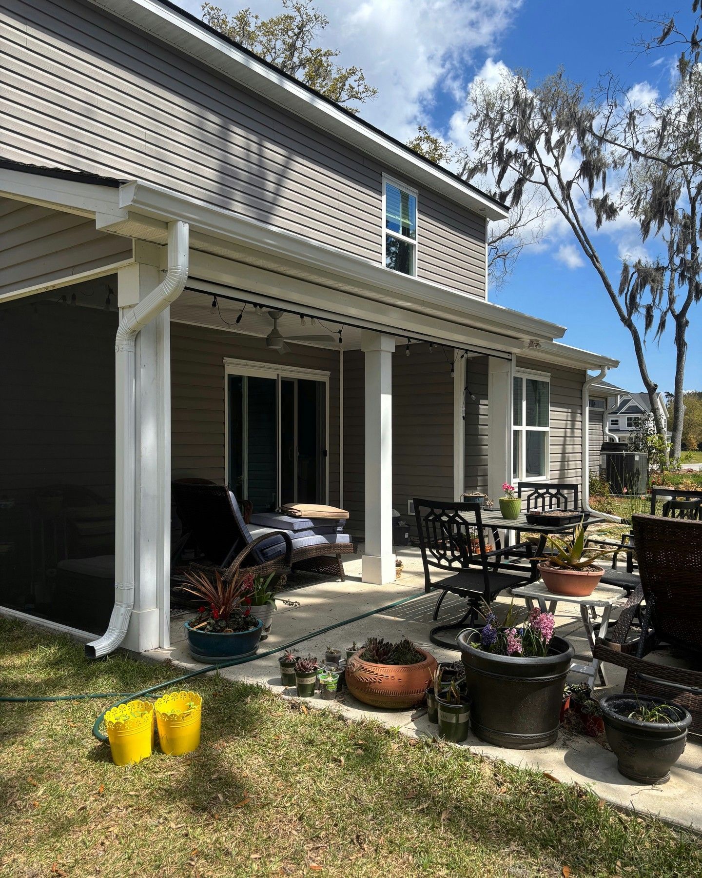 A grey house with a covered patio, outdoor furniture, and several potted plants on a concrete slab near a grassy yard.