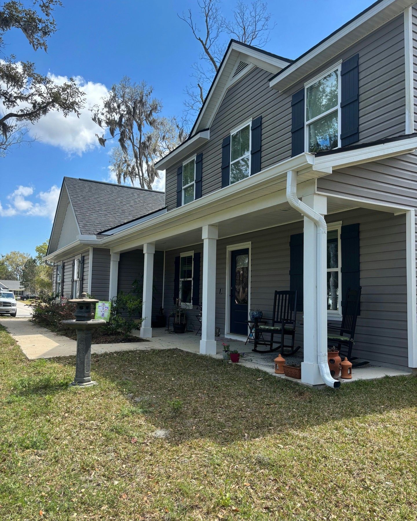 A two-story grey-sided house with a white wraparound porch, black shutters, and a birdbath in the front yard.