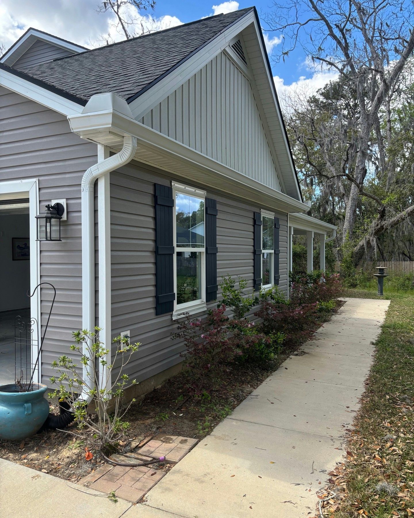 A side view of a gray-sided home with a white downspout, shuttered windows, and a concrete path leading into the yard.