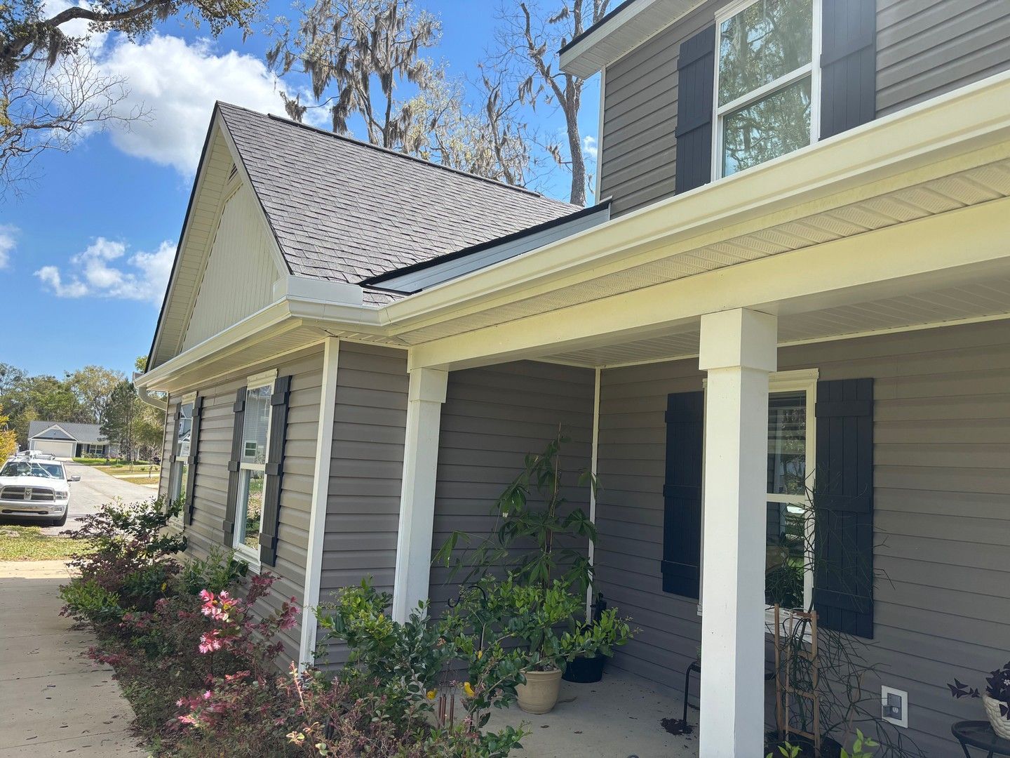 A gray house with white trim, dark shutters, and a porch on a sunny day with trees in the background.