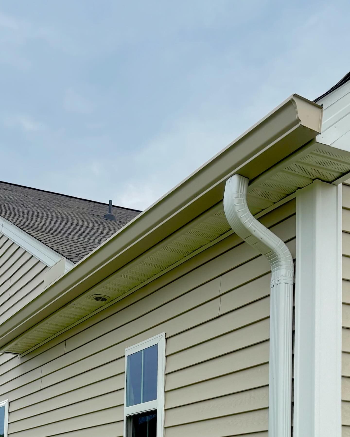 Tan siding on a house exterior featuring a roof overhang with a tan gutter and a white downspout against a blue sky.