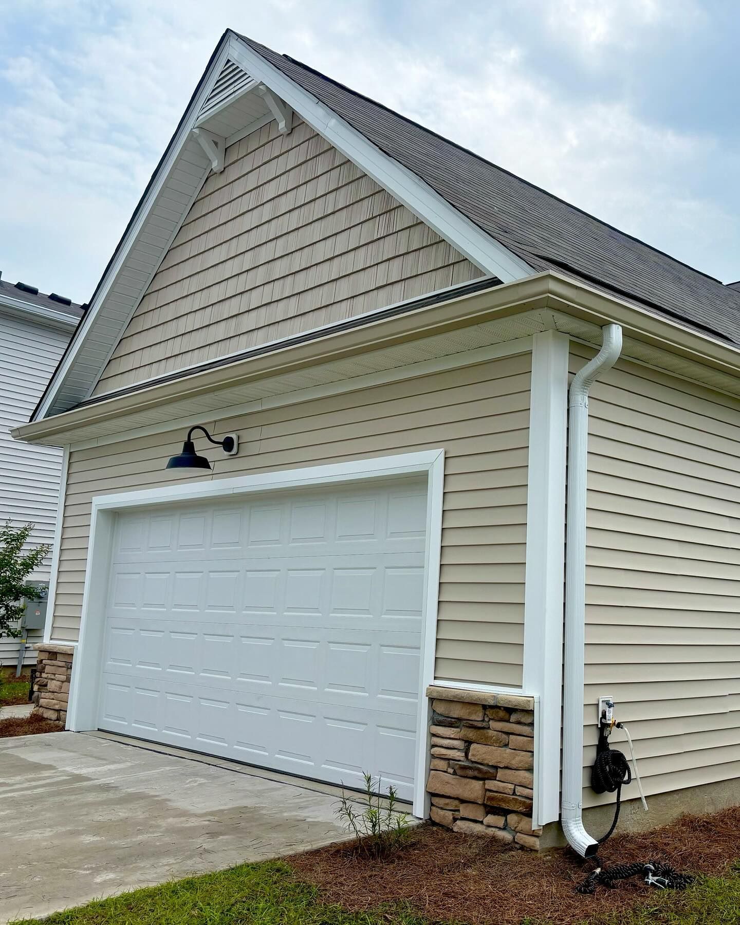 A tan garage with horizontal siding, stone base corners, a white door, a black light fixture, and a white downspout.