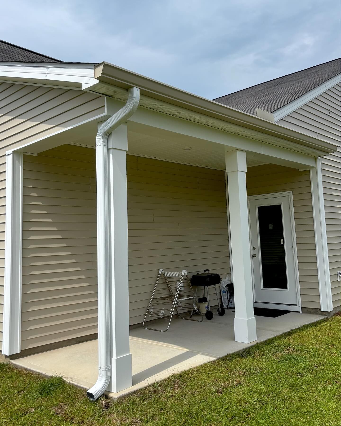 A tan-sided house with a covered concrete patio, a white door, and a white downspout running along a structural post.