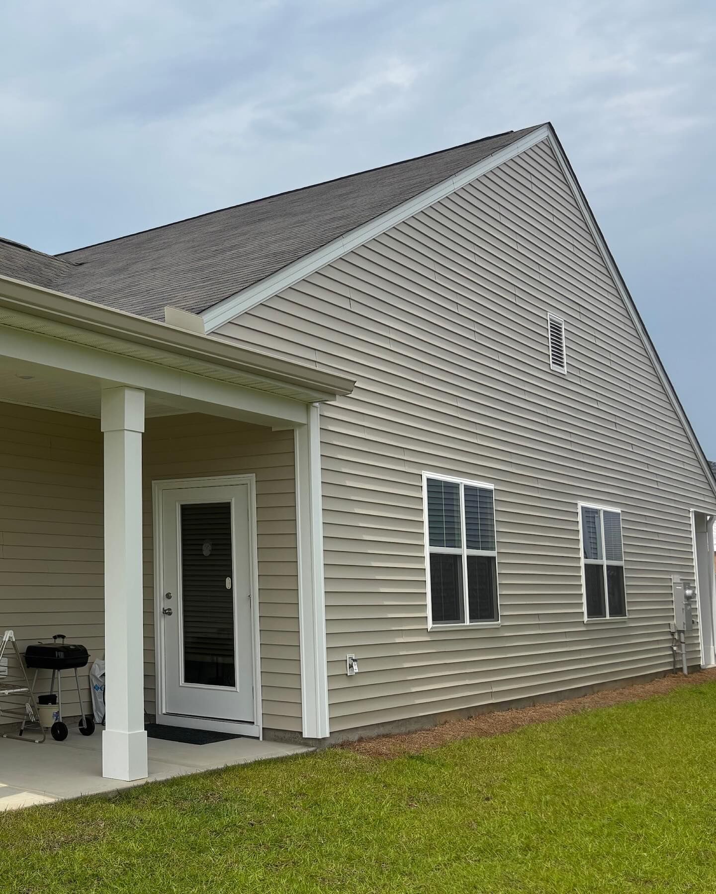 Tan-sided house exterior with a covered patio, two windows, and a grassy lawn under a cloudy sky.