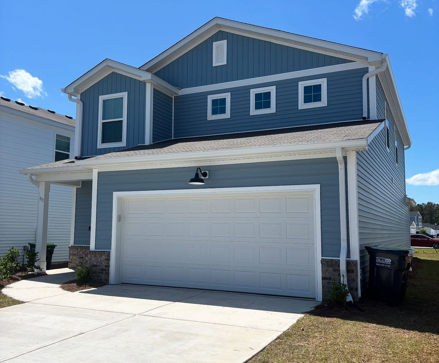 A two-story, blue-sided suburban house with a white two-car garage, stone trim, and a concrete driveway under a blue sky.