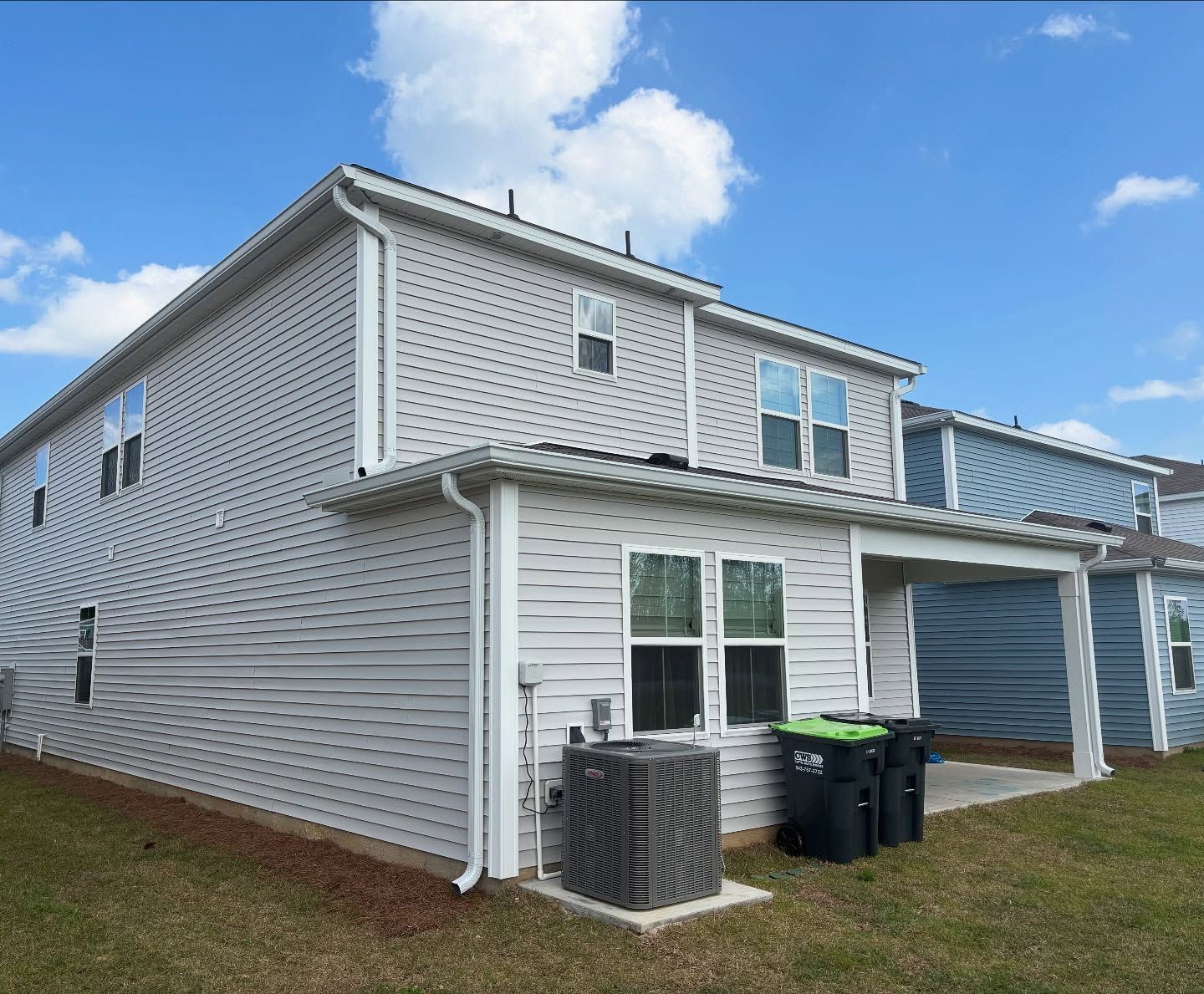 Back view of a two-story light gray vinyl-sided house with a central air conditioning unit and two trash bins.