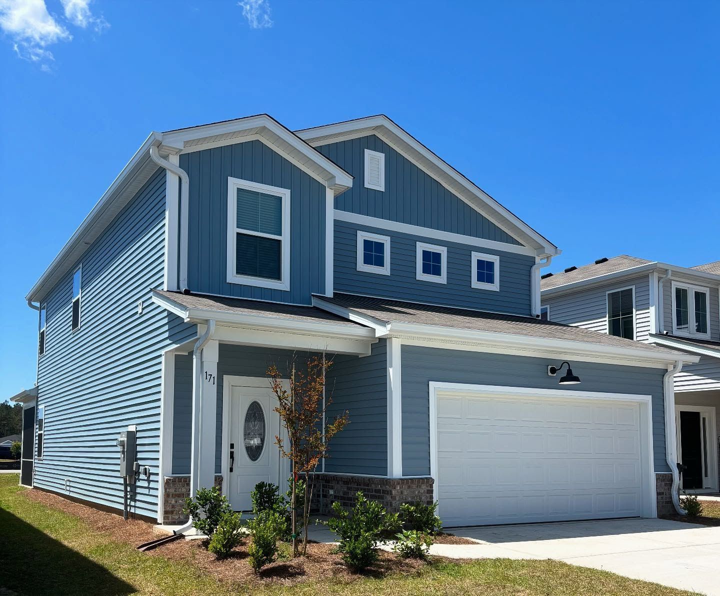 A two-story, blue-sided suburban house with a white front door and an attached two-car garage under a clear blue sky.