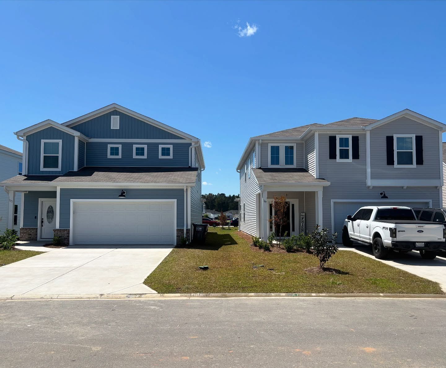 Two suburban homes side-by-side, one blue and one light gray, under a clear blue sky, with a white truck in the driveway.