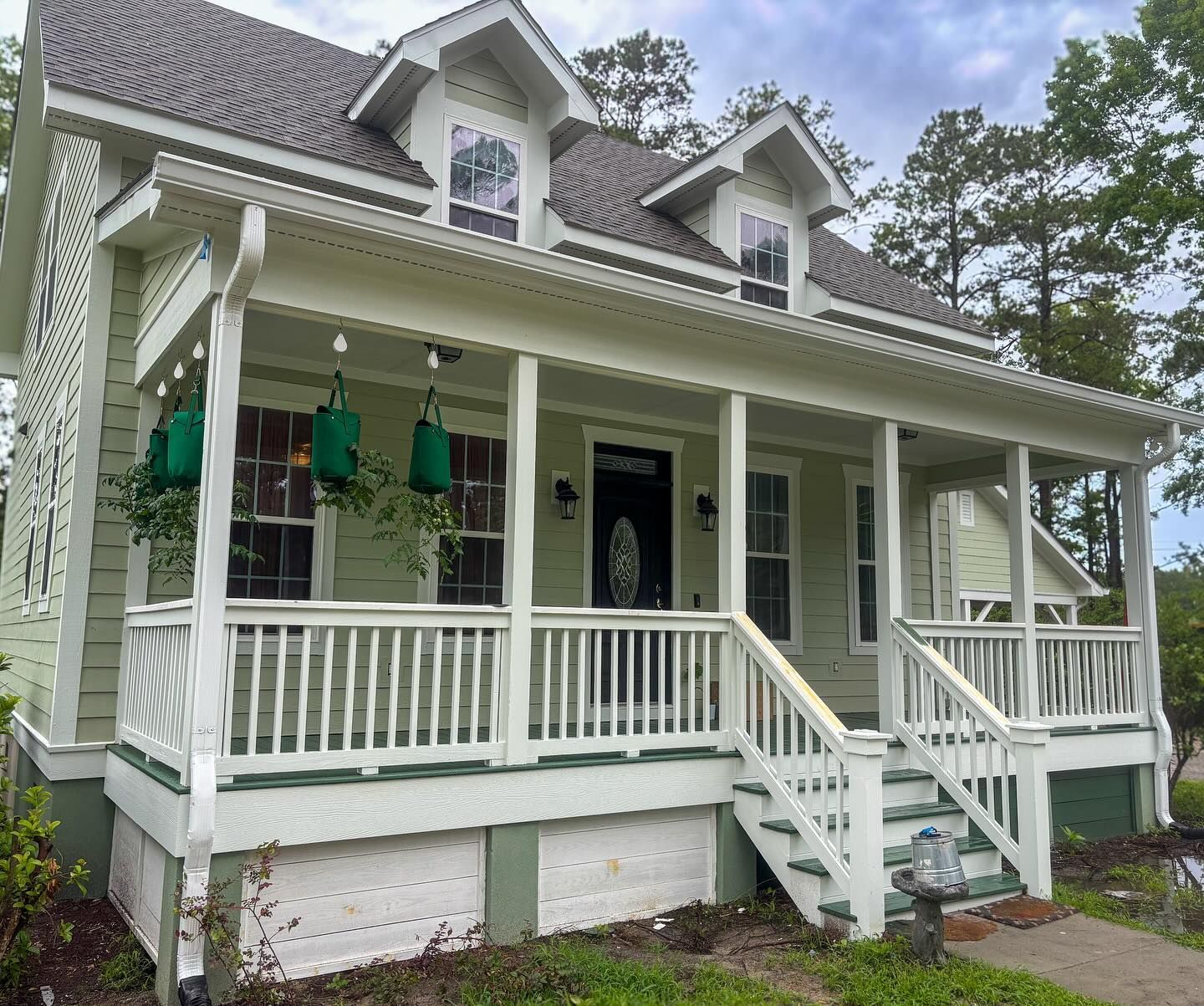 A light green, two-story farmhouse with a white wraparound porch, dark shutters, and two dormer windows.