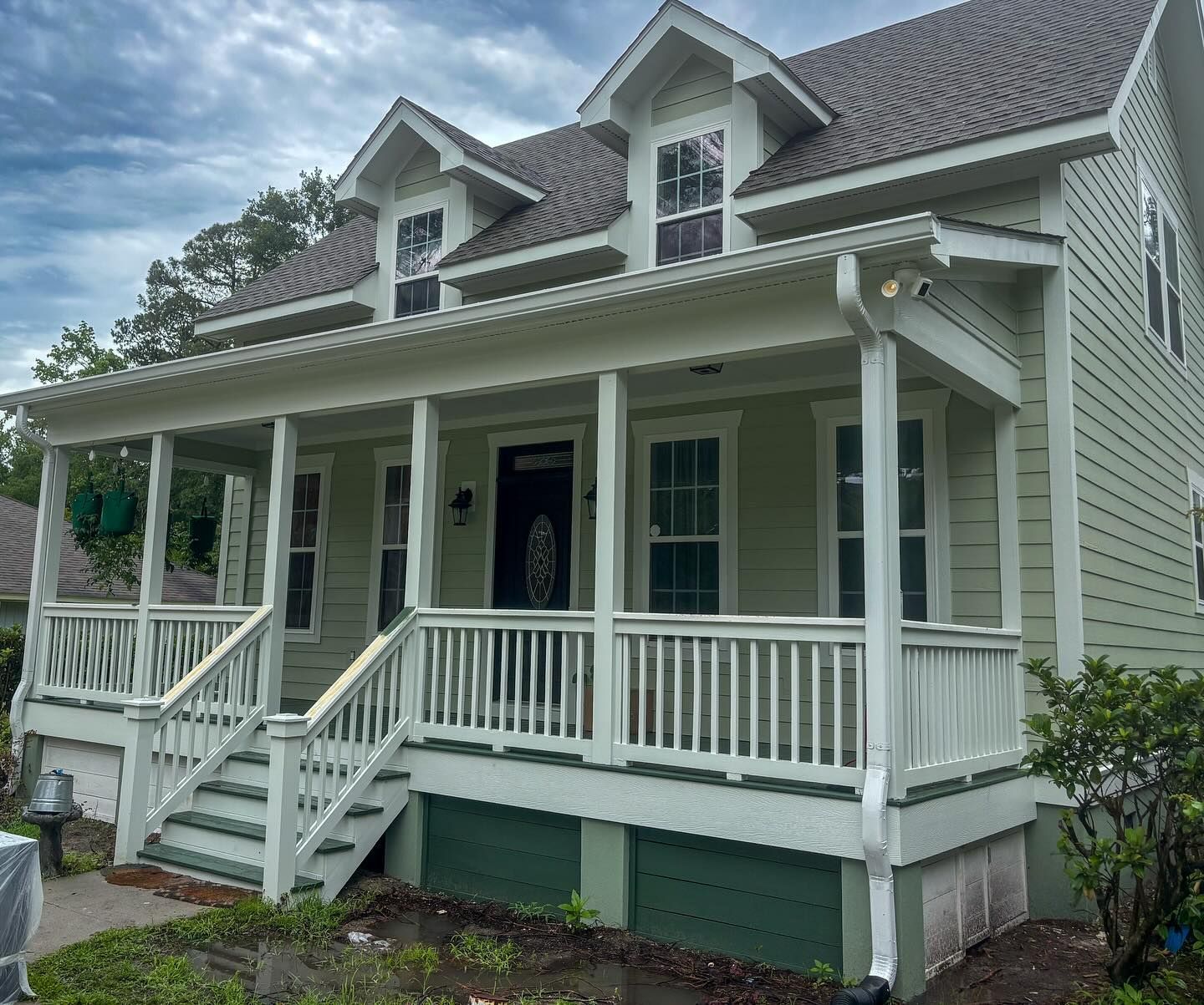 Light green two-story house with a white wraparound porch, dark door, and two dormer windows under a cloudy sky.