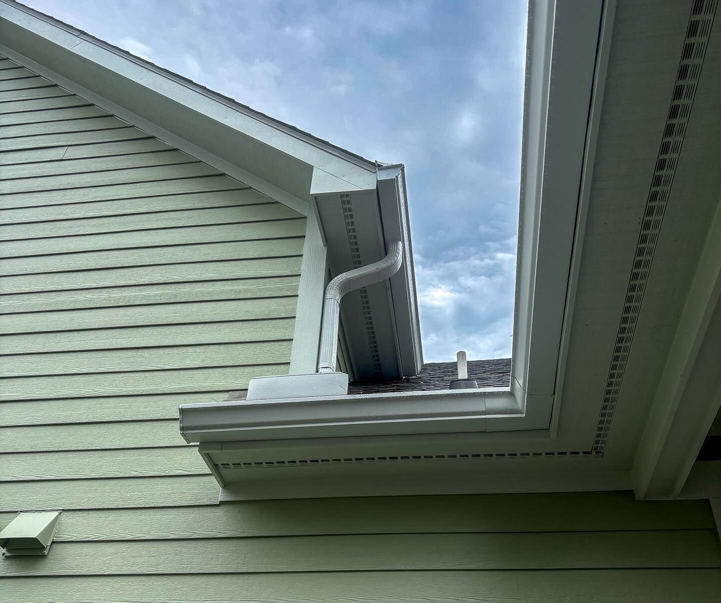 A low-angle view of a house corner showing light green siding, white gutters, a downspout, and a soffit against a blue sky.