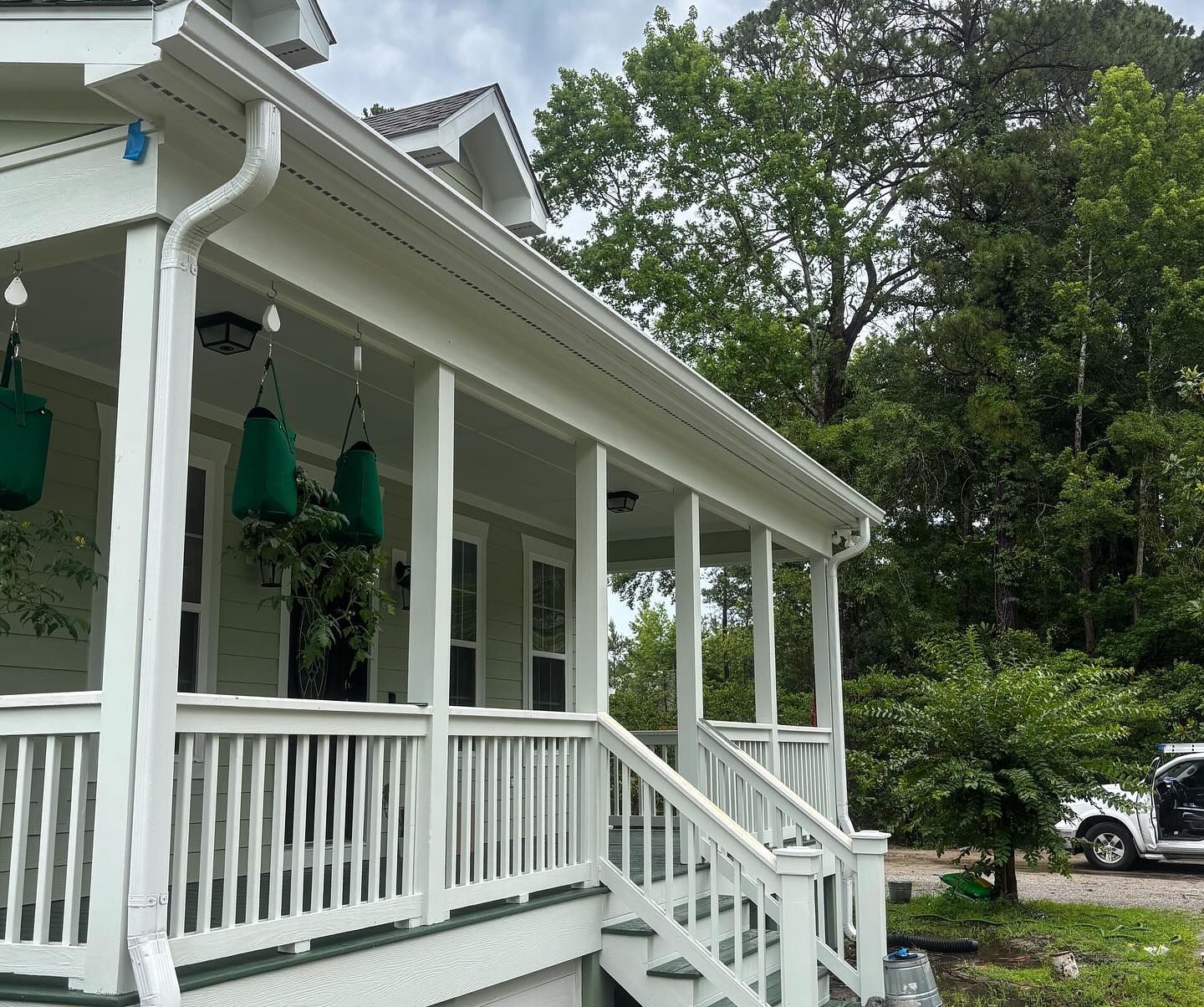 A white wraparound porch on a light green house with green hanging baskets, a white railing, and stairs leading down.