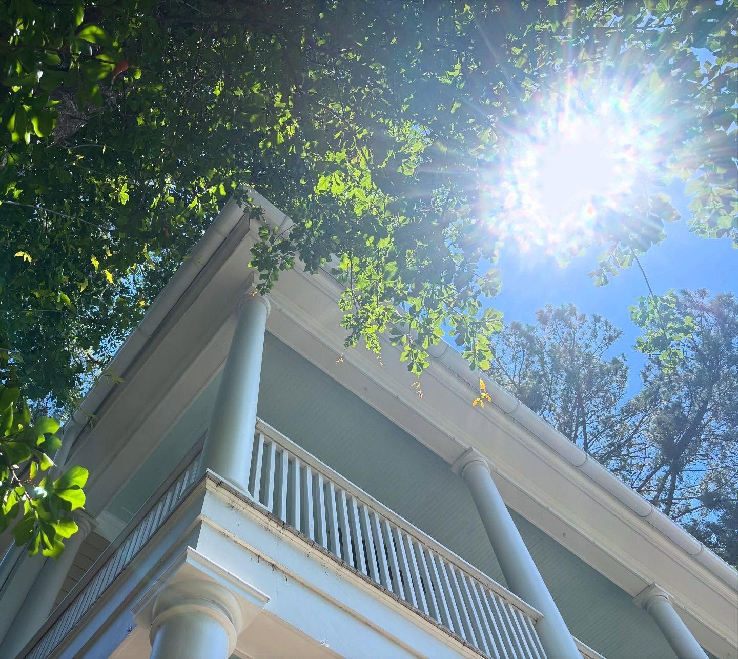 A low-angle view of a blue, two-story house with tall white columns and a balcony, framed by sunlit green tree leaves.