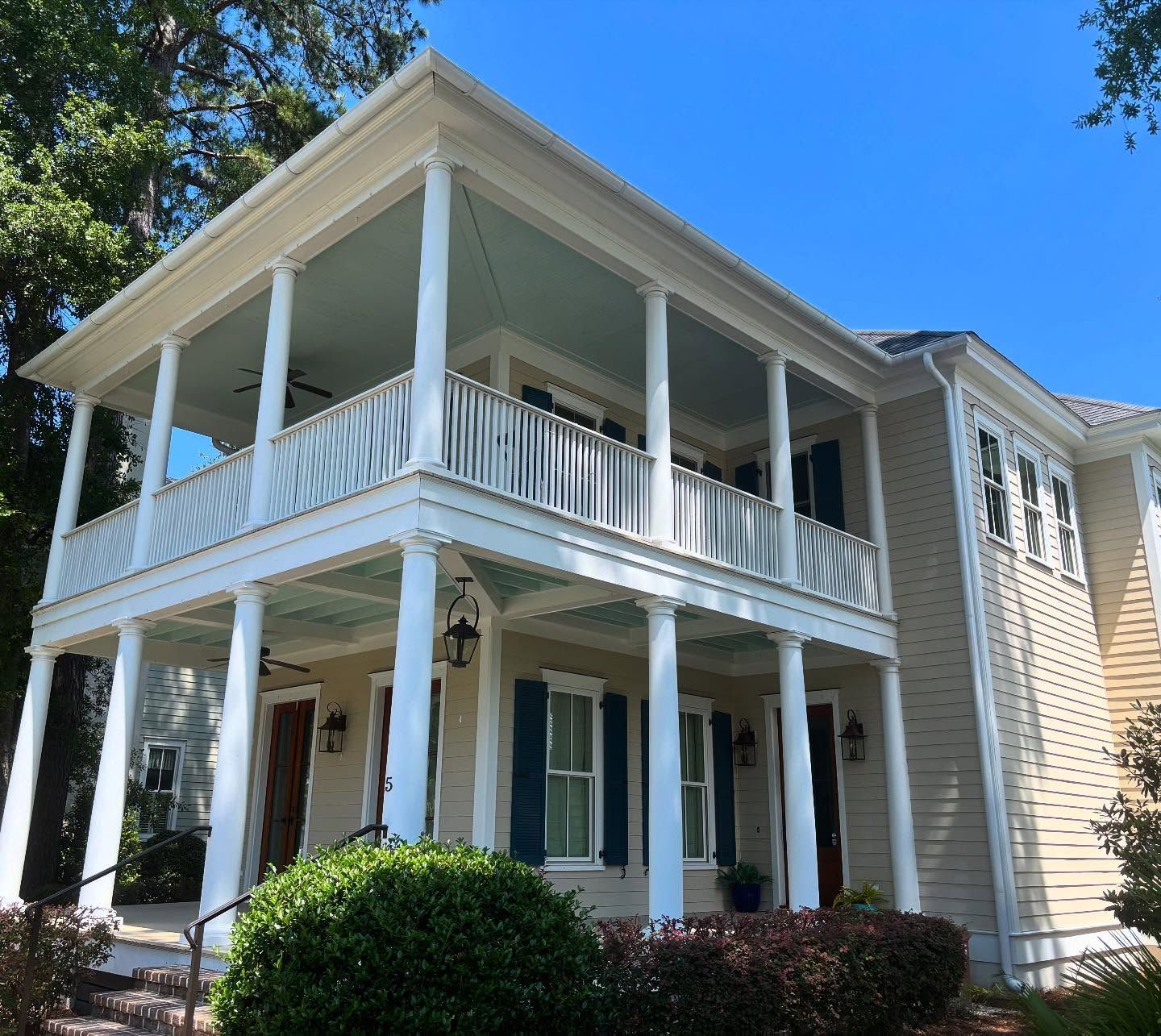 A beige, two-story house with white columns, a wrap-around porch, and blue shutters, viewed from a low angle.