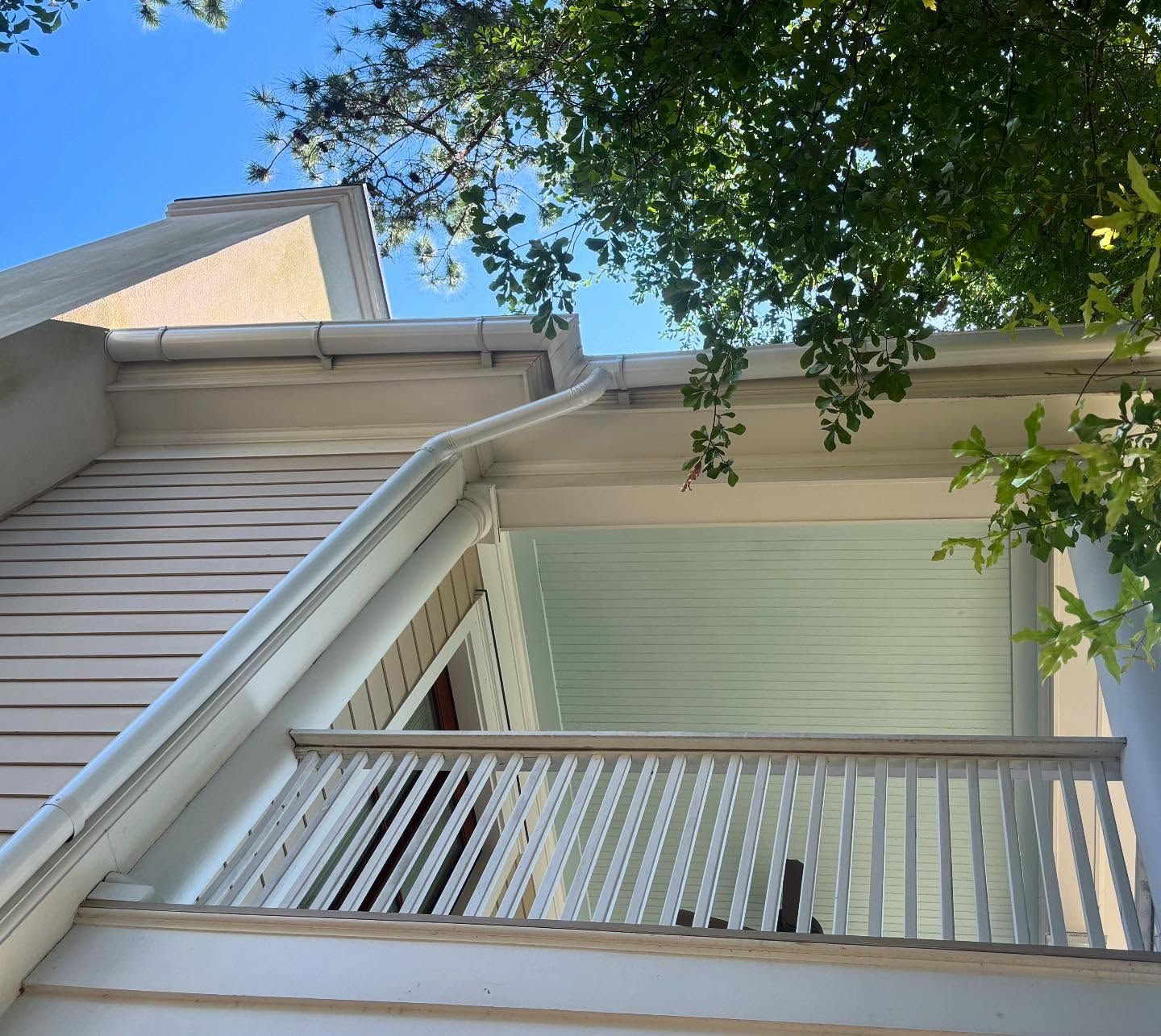 A low-angle view of a white, multi-story building exterior with a balcony, siding, gutters, and green tree branches.