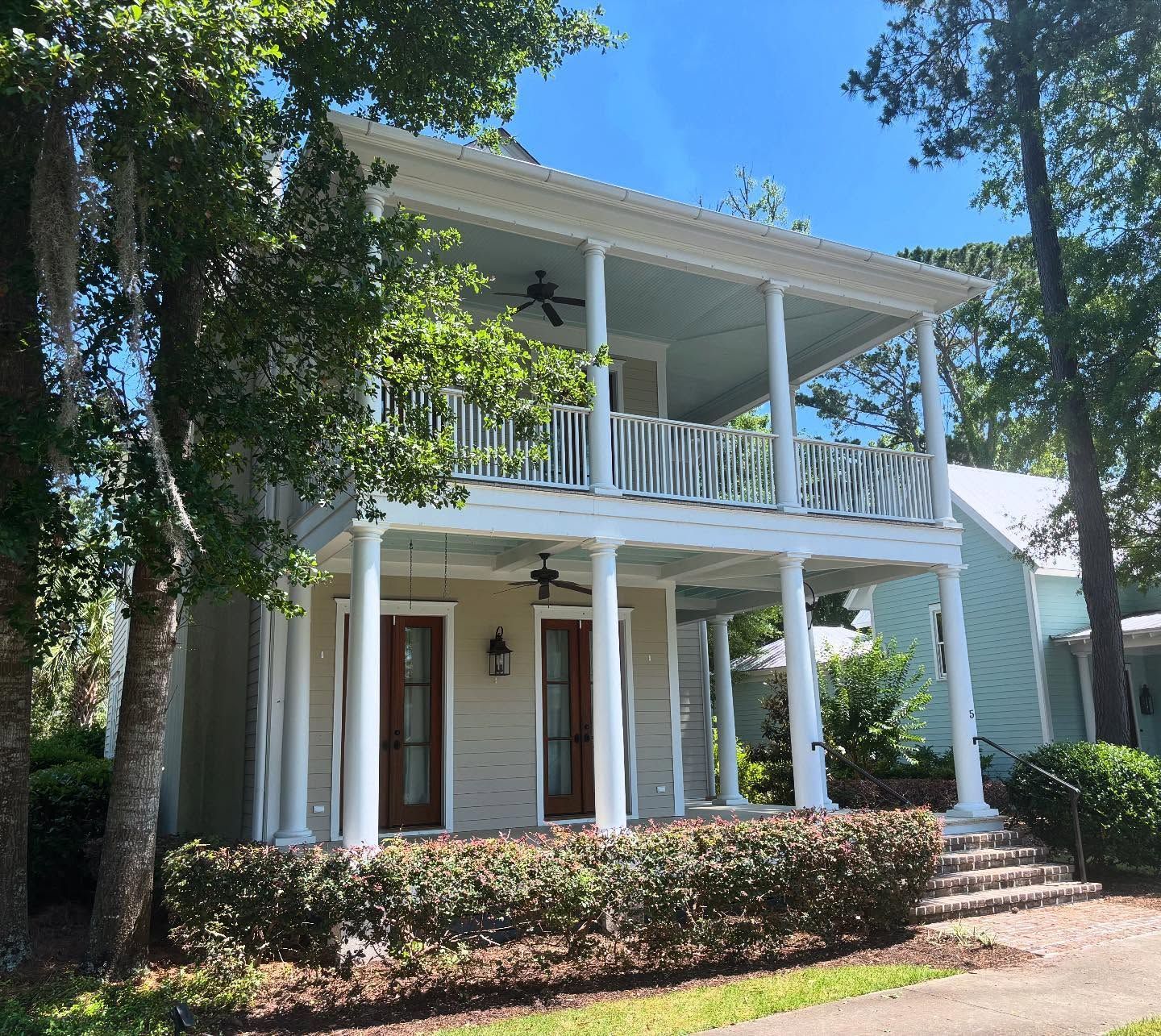 A two-story house with a large white porch, columns, wooden doors, and light tan siding, framed by green trees.