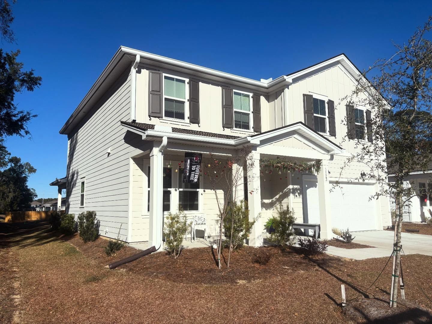 A two-story, cream-colored suburban house with dark shutters, a covered porch, and a side-load garage under a blue sky.