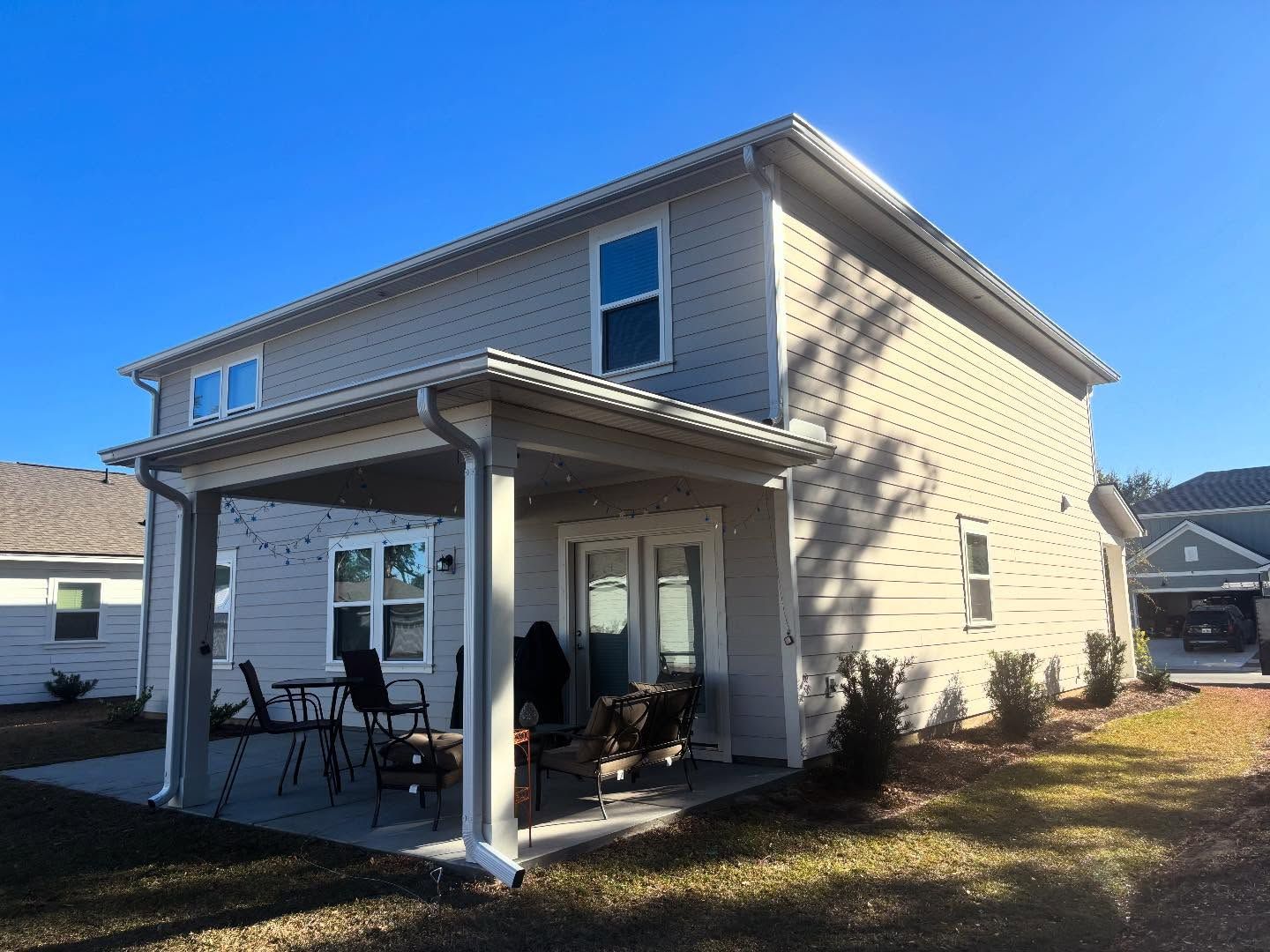 Beige two-story house with a covered back patio, patio furniture, and string lights on a sunny day.