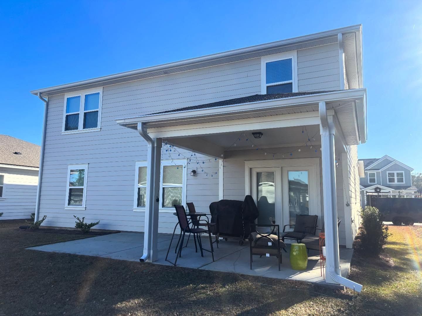 A light-gray two-story house with a covered back patio, patio furniture, and a concrete slab under a bright blue sky.
