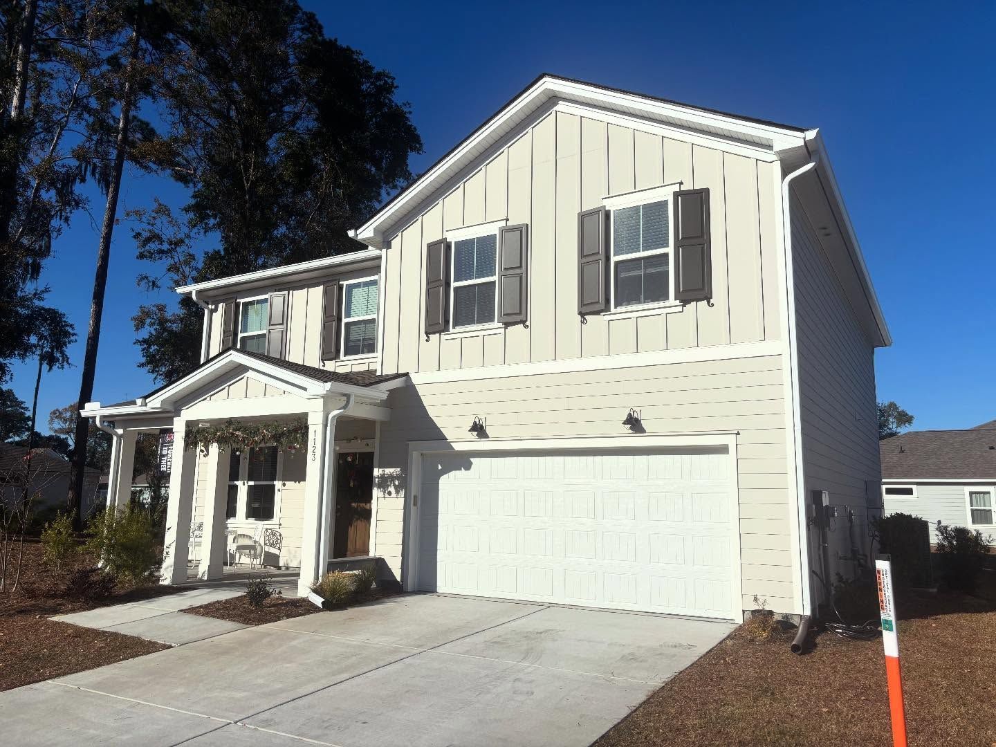 A two-story, light-colored suburban house with a white garage, covered front porch, and blue sky.